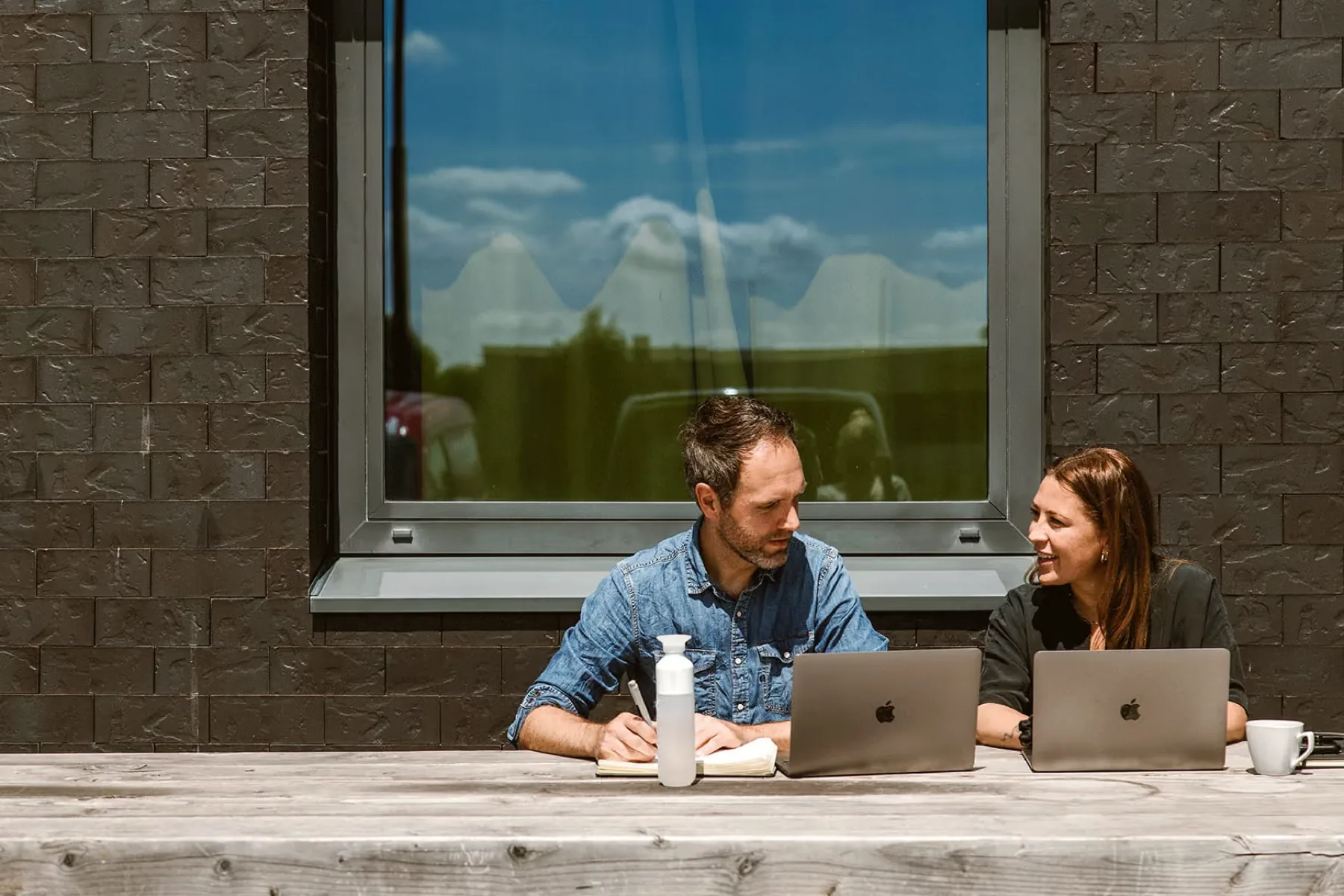 Twee collega's werken buiten aan een houten tafel met hun laptops in de zon.